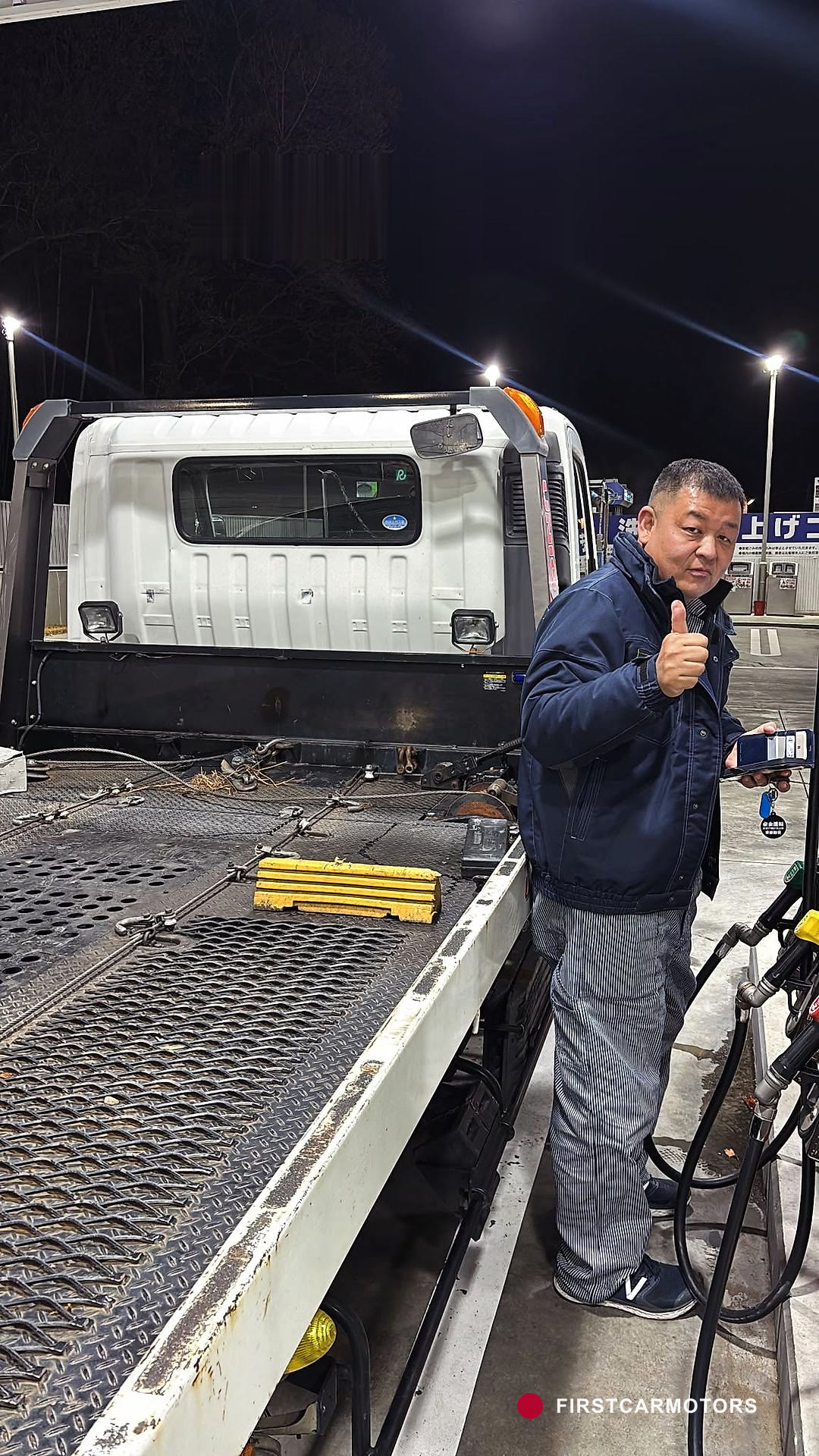 Amanuel steht lächelnd mit erhobenem Daumen neben einem japanischen Kollegen an einer Tankstelle vor dem beladenen Transporter.