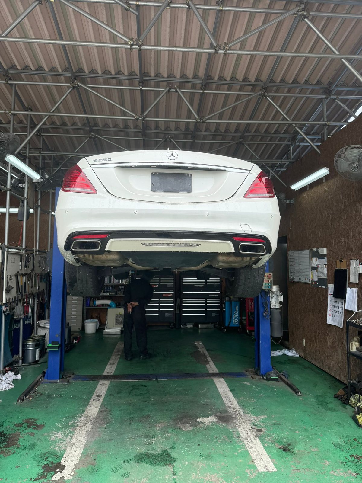 Ein weißer Mercedes steht angehoben auf einer Zweisäulen-Hebebühne in einer Werkstatt, darunter arbeitet ein Mechaniker.